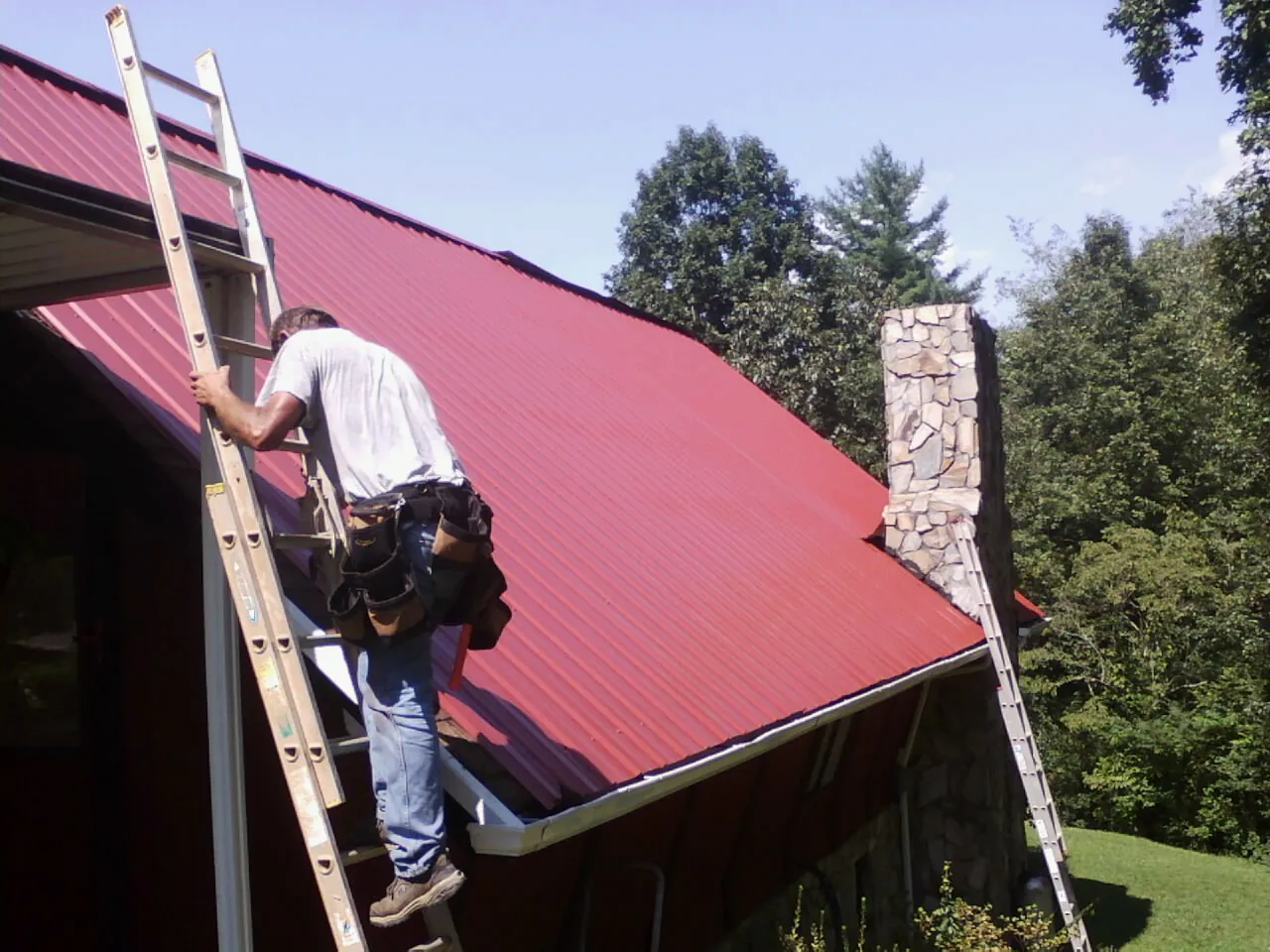 Roofer on a ladder beside a red metal roof and stone chimney — Soffit & Fascia Repair by Roofing Brothers in Moon